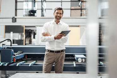 Lächelnder reifer Geschäftsmann, der ein digitales Tablet hält, während er vor einer Maschine in einer Fabrik steht - JOSEF02316