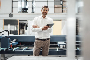 Smiling mature businessman holding digital tablet while standing against machinery in factory - JOSEF02316