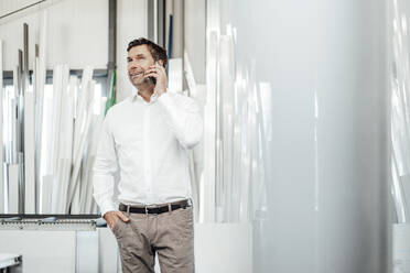 Männlicher Manager, der mit dem Smartphone spricht, während er mit der Hand in der Tasche in der Industrie steht - JOSEF02317