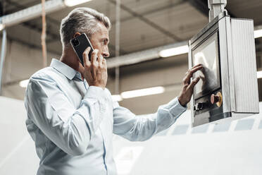 Älterer männlicher Ingenieur, der mit einem Mobiltelefon spricht, während er eine Anlage in einer Fabrik bedient - JOSEF02334