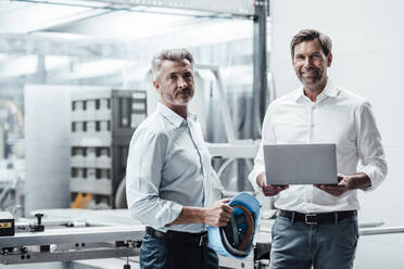 Lächelnder männlicher Ingenieur mit Laptop, der neben einem Kollegen mit Schutzhelm in einer Fabrik steht - JOSEF02340