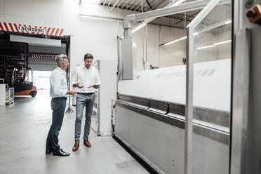 Male engineers discussing over digital tablet while standing by machinery in industry - JOSEF02372