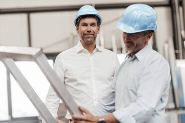 Male engineer smiling while colleague holding metallic frame in industry - JOSEF02386