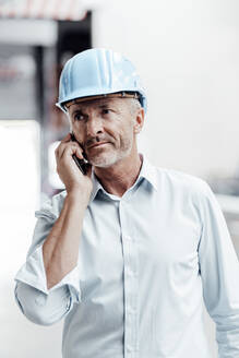 Männlicher Ingenieur mit Schutzhelm schaut weg, während er mit einem Smartphone in der Industrie spricht - JOSEF02390