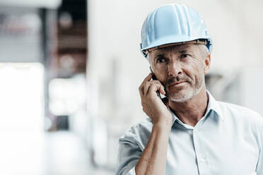 Mature male engineer in hardhat looking away while talking on mobile phone in factory - JOSEF02391