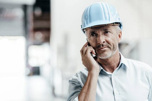 Reifer männlicher Ingenieur mit Schutzhelm, der wegschaut, während er in einer Fabrik mit einem Mobiltelefon spricht - JOSEF02391