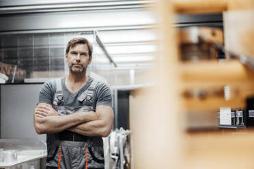 Confident mature male worker standing with arms crossed in factory - JOSEF02421