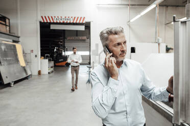 Businessman talking on mobile phone while colleague walking on background at industry - JOSEF02430