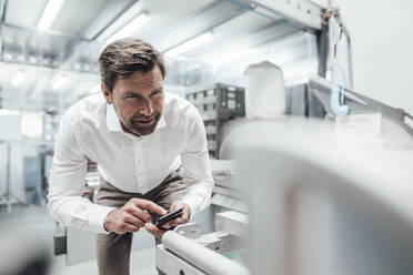 Male engineer using smart phone while examining manufacturing equipment in industry - JOSEF02442