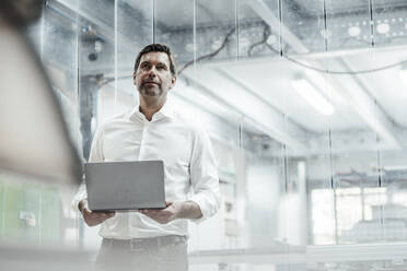 Thoughtful male engineer with laptop looking away while standing in industry - JOSEF02450