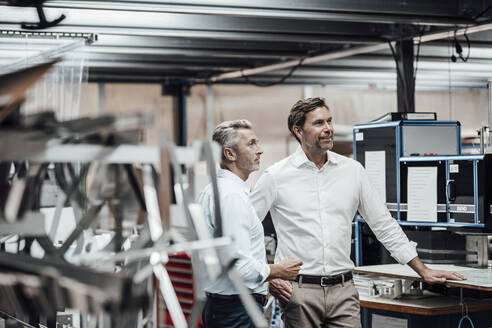 Ein reifer Kollege diskutiert mit einem lächelnden Manager, während er an einer Fertigungsmaschine in einer Fabrik steht - JOSEF02459