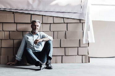 Thoughtful businessman sitting against boxes in factory - JOSEF02468