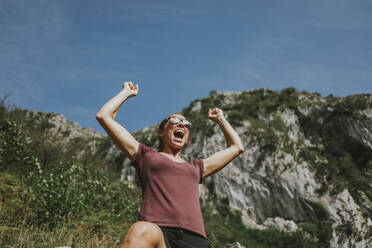 Smiling female trekker celebrating victory with arms raised shouting against sky on sunny day - DMGF00270