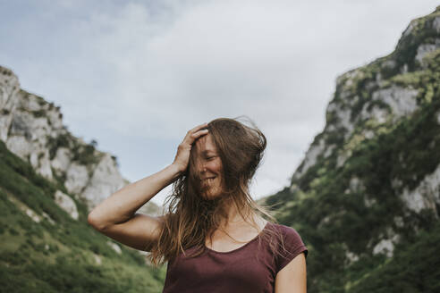 Happy female trekker with tousled long brown hair against mountain range - DMGF00276