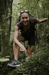 Smiling female trekker bending over by plants in forest - DMGF00288