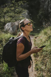 Frau schaut weg, während sie ihr Smartphone zur Navigation beim Wandern an einem sonnigen Tag hält - DMGF00296