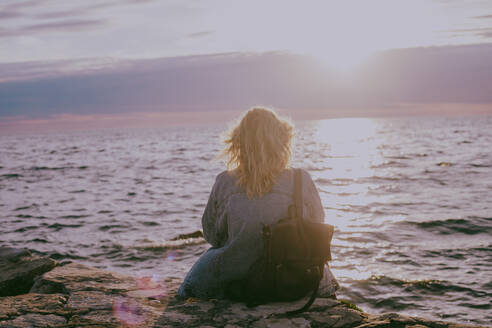 Rückenansicht einer blonden Frau, die bei Sonnenuntergang am Meer sitzt - CAVF90330
