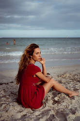Nachdenkliche Frau, die wegschaut, während sie am Ufer des Strandes sitzt - MAUF03573