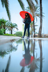 Frau mit einer Maske und einem roten Regenschirm, der sich auf einer Pfütze spiegelt - CAVF90570