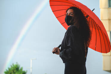 Frau mit einer Maske und einem roten Regenschirm steht in der Nähe eines Regenbogens - CAVF90571