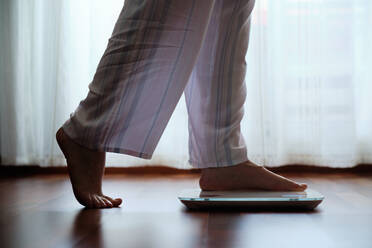 Barfüßige, gesichtslose Frau in gemütlichem Pyjama, die morgens auf einer digitalen Gewichts- und Körperfettwaage mit Anzeige auf dem Badezimmerboden steht - ADSF17155