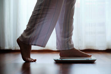 Crop faceless barefoot female in cozy pajama standing on digital weight and body fat scales with display on bathroom floor in the morning - ADSF17155