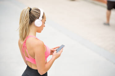 Side view of slender sportswoman in sportswear listening to music in headset while surfing internet on cellphone and standing on embankment near palms and sea - ADSF17158