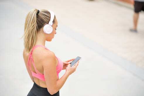 Seitenansicht einer schlanken Sportlerin in Sportkleidung, die mit einem Headset Musik hört, während sie mit ihrem Handy im Internet surft und auf einem Damm in der Nähe von Palmen und Meer steht - ADSF17158
