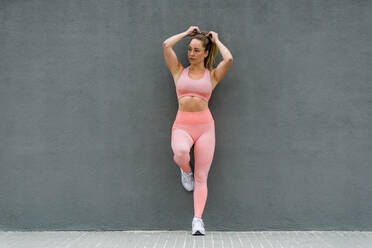 Ganzkörper einer fitten Sportlerin in Sportkleidung und Turnschuhen, die auf einem Bein angelehnt an der Böschung nahe der grauen Wand steht und wegschaut - ADSF17159