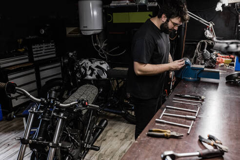 Seitenansicht eines konzentrierten männlichen Mechanikers mit Tätowierungen am Schraubstock und Reparatur einer roten Rückleuchte eines Motorrads bei der Arbeit in der Werkstatt - ADSF17166