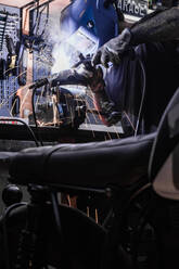 Seitenansicht eines anonymen männlichen Arbeiters mit Schutzhelm, der an der Werkbank steht und Metalldetails mit einem Schweißwerkzeug befestigt - ADSF17169