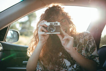Zufriedene Frau in Freizeitkleidung und mit Brille im Auto sitzend beim Fotografieren mit Digitalkamera im Sonnenlicht bei Gegenlicht - ADSF17175