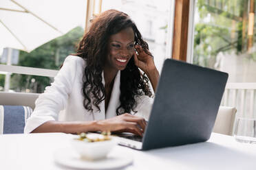 Elegant lächelnde afrikanisch-amerikanische Geschäftsfrau in weißer Jacke beim Surfen am Laptop weiß, die aus der Ferne im Café arbeitet - ADSF17180