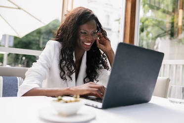 Elegant smiling African America businesswoman in white jacket browsing laptop white working remotely in cafe - ADSF17180