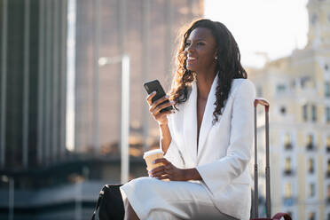 Positive African American businesswoman in elegant white clothes browsing cellphone and having coffee while sitting on street with suitcase - ADSF17182