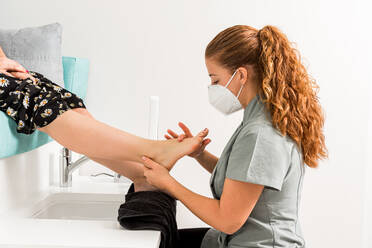 Cropped unrecognizable female client sitting on chair and getting pedicure treatment from professional master in protective mask in modern beauty salon - ADSF17198