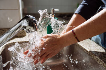 Nicht erkennbare Person, die sich die Hände mit Wasser reinigt, während sie in der Nähe des Waschbeckens im vom Sonnenlicht beleuchteten Badezimmer steht - ADSF17207