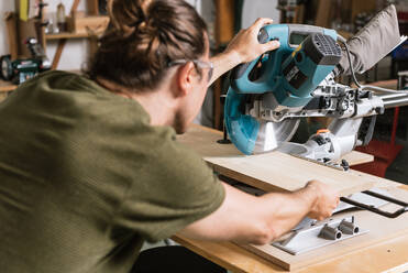 Back view of male carpenter in protective goggles cutting wooden plank with miter saw while working in bright workshop - ADSF17209