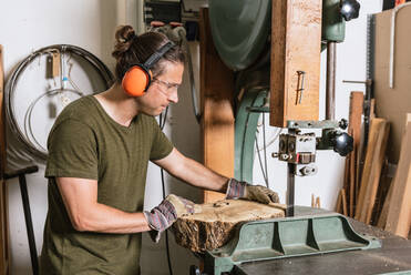 Focused male carpenter in protective ear muffs and goggles cutting piece of wood with band saw in shabby workshop - ADSF17211