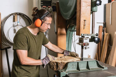 Fokussierter männlicher Schreiner mit Gehörschutzkapseln und Schutzbrille schneidet Holzstück mit Bandsäge in schäbiger Werkstatt - ADSF17211