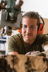 Focused male carpenter in protective ear muffs and goggles cutting piece of wood with band saw in shabby workshop - ADSF17212