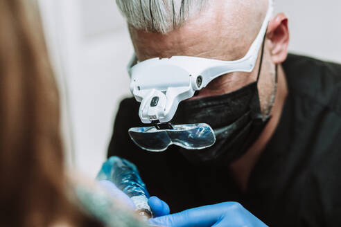 Geschäftiger männlicher Tätowierer mit Handschuhen und Maske, der eine Maschine mit Tinte benutzt und ein Bild auf dem Arm eines Kunden im Salon erstellt - ADSF17218