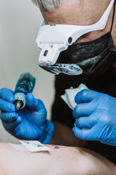 Geschäftiger männlicher Tätowierer mit Handschuhen und Maske, der eine Maschine mit Tinte benutzt und ein Bild auf dem Arm eines Kunden im Salon erstellt - ADSF17219