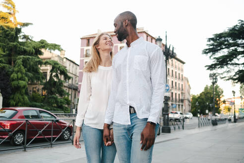 Glücklich lächelndes gemischtrassiges Paar in Jeans und weißem Hemd, das bei Sonnenschein auf dem Bürgersteig spazieren geht und in der Stadt Händchen hält - ADSF17235