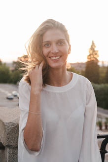 Erfreute positive Frau in weißer Bluse ruht auf Straßengeländer und schaut glücklich in die Kamera während des Sonnenuntergangs - ADSF17240