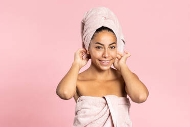 Fröhliche junge ethnische Frau mit nackten Schultern, Badetuch auf dem Kopf und perfekter olivfarbener Haut schaut in die Kamera vor rosa Hintergrund, während sie Schönheit und Hautpflegekonzept darstellt - ADSF17251