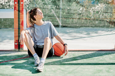 Seitenansicht einer jungen sportlichen Frau mit Basketball, die am Boden auf dem Platz sitzt und sich nach dem Training an einem sonnigen Sommertag entspannt - ADSF17264