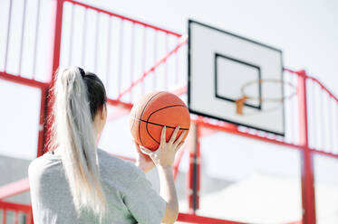 Back view of young sporty woman performing shot while playing basketball alone on sports ground - ADSF17265