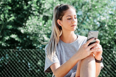 Ernte einer jungen Frau in Sportkleidung, die an einem sonnigen Sommertag in einem Mobiltelefon surft, während sie sich nach dem Training auf einem Sportplatz ausruht - ADSF17266