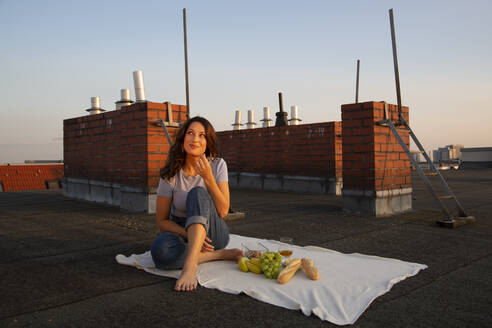 Thoughtful young woman having fruits during picnic on rooftop - NGF00704